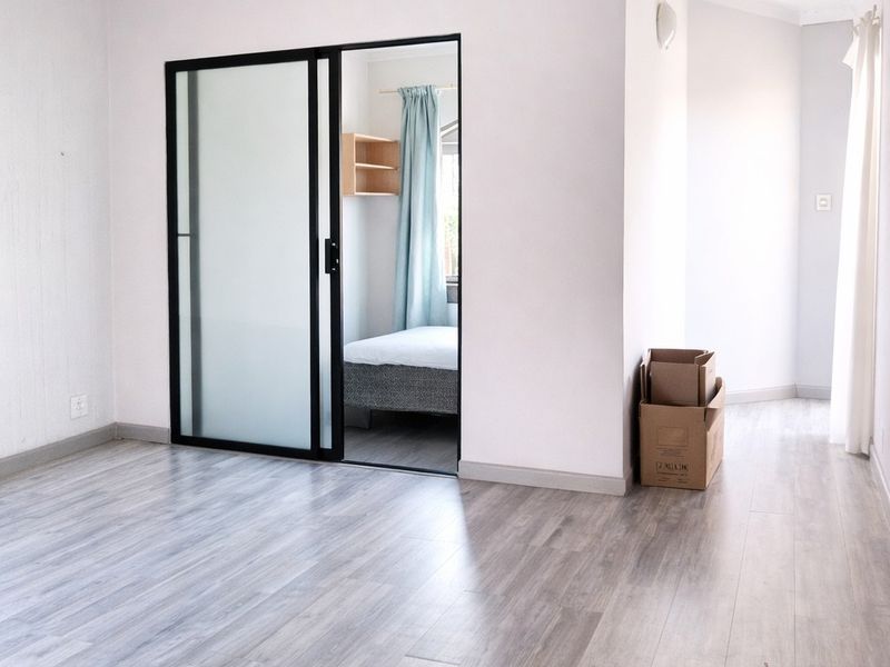 A bright, empty living area featuring modern grey wood-look flooring and a sleek black-framed sliding glass door leading to an adjacent bedroom.