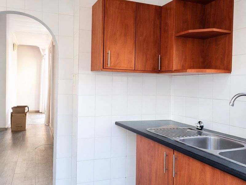 A compact kitchen area featuring wooden cabinetry, a dark laminate countertop, and a stainless steel sink, adjacent to an arched hallway entrance.