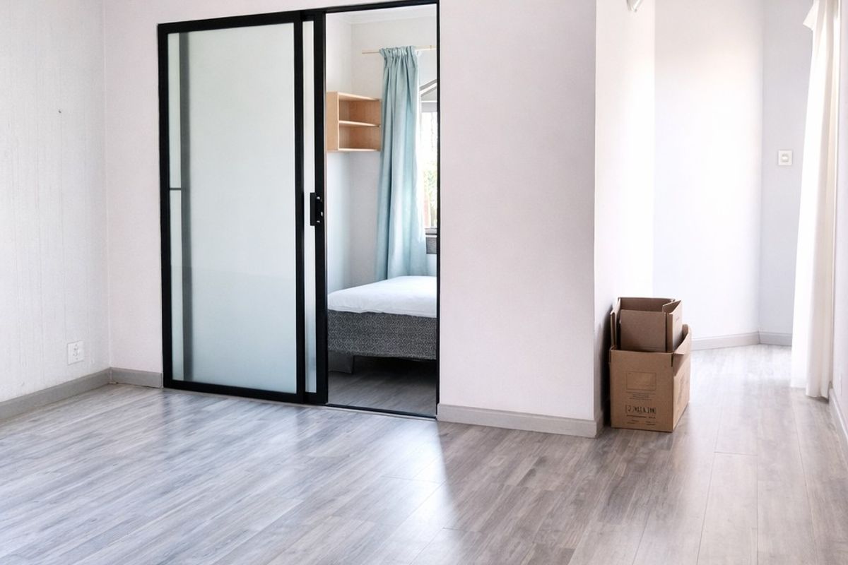A bright, empty living area featuring modern grey wood-look flooring and a sleek black-framed sliding glass door leading to an adjacent bedroom.
