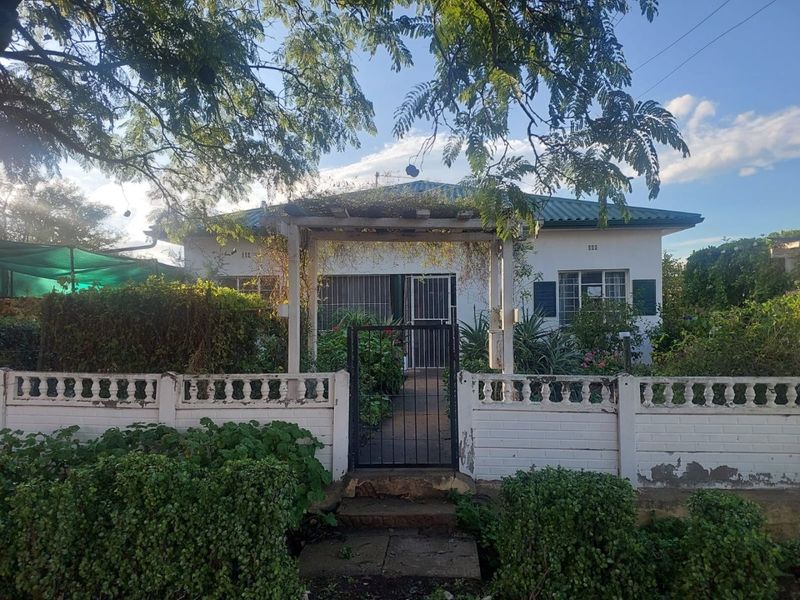 Front view of a single-story house featuring a white facade, metal roof, and a gated entrance with a timber pergola structure.