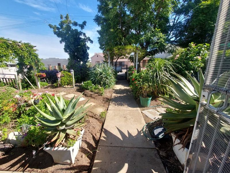A landscaped front garden featuring a concrete pathway leading to a gated entrance, surrounded by established shrubs, trees, and garden beds.