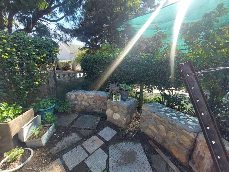 A rustic garden area featuring stone-built seating benches, paved pathways, and a shaded canopy structure.