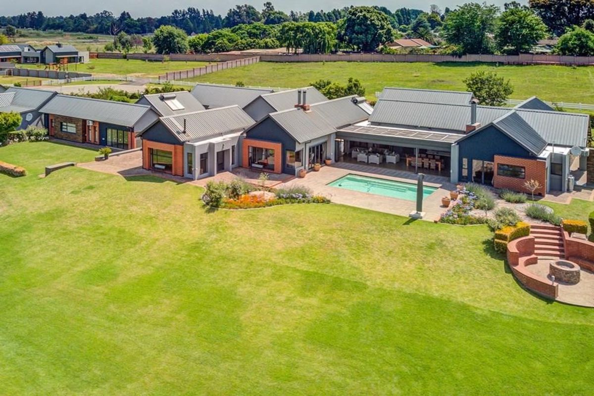 Aerial view of a sprawling modern farmhouse with a swimming pool, covered patio, landscaped gardens, and a brick-built fire pit area.