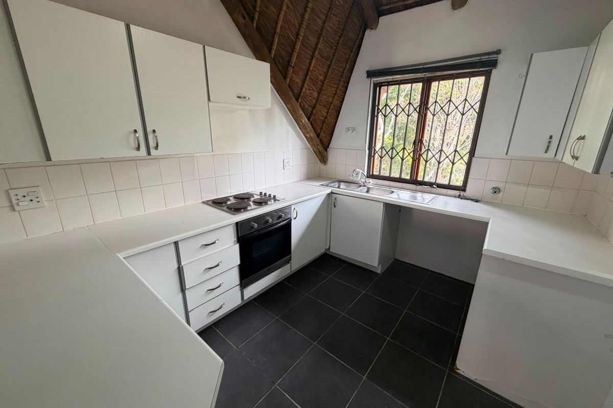 A functional kitchen featuring white cabinetry, built-in oven and hob, tiled countertops, and dark floor tiles, set under a unique thatched roof structure.