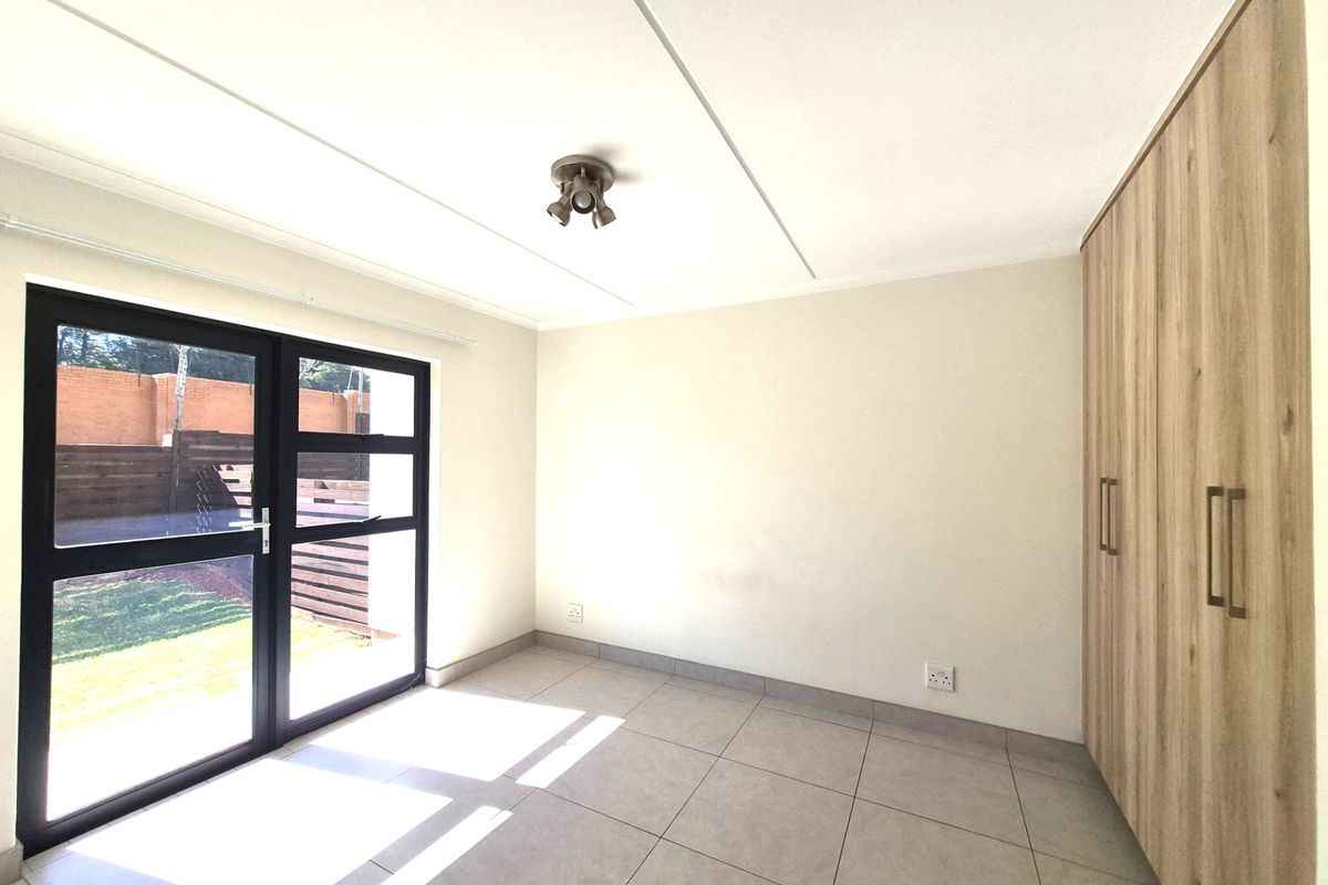A bright, empty bedroom featuring neutral floor tiles, built-in wooden cupboards, and a glass door providing direct access to the garden.