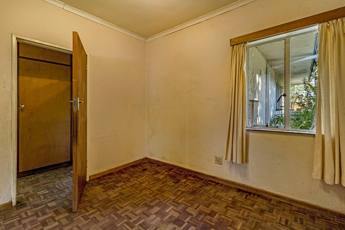 An empty bedroom featuring original parquet flooring, a built-in cupboard, and a window overlooking the exterior area, requiring cosmetic updates.