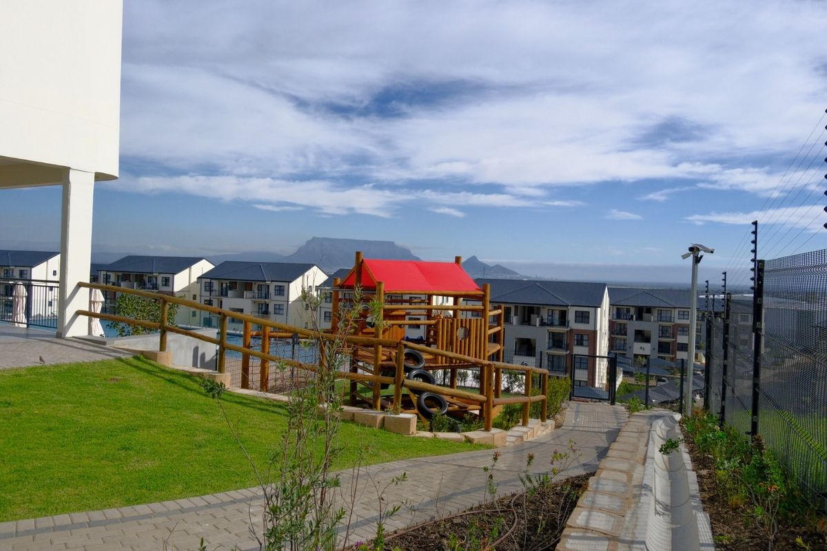 Communal outdoor area featuring a paved walkway, landscaped lawn, and a children's play structure, set within a secure apartment complex with mountain views.