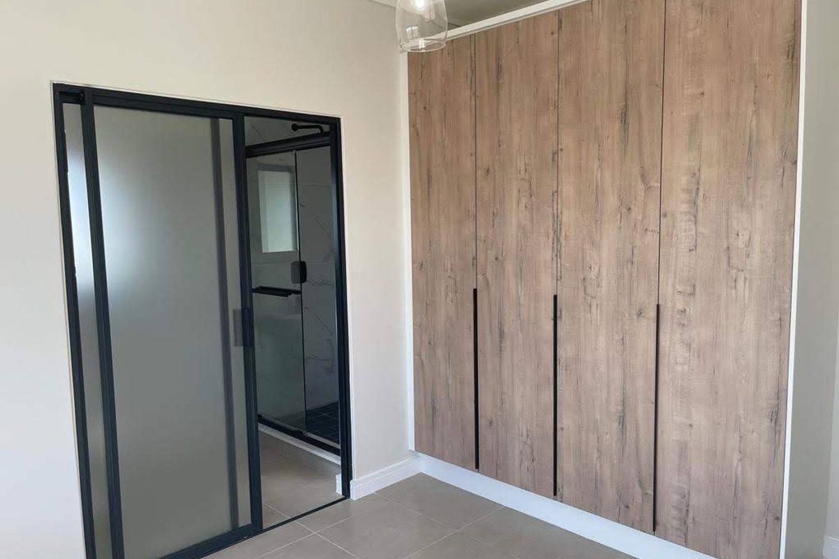 A modern bedroom featuring floor-to-ceiling wood-grain built-in cupboards and a sleek black-framed glass door leading to an en-suite bathroom.