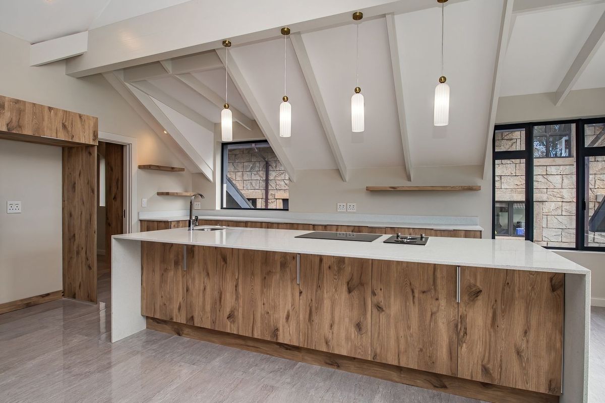 Modern kitchen featuring a large island with stone countertops, wood-grain cabinetry, built-in oven, and exposed vaulted ceiling beams.