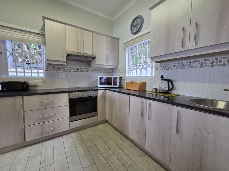 A well-maintained kitchen featuring light wood-grain cabinetry, granite countertops, built-in oven, electric hob, and tiled splashbacks with security bars on the windows.