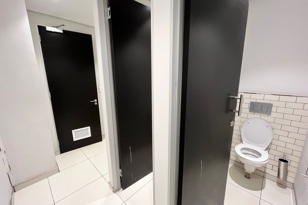 A modern office restroom featuring tiled flooring, subway tile backsplash, and wall-mounted toilet fixtures with black privacy doors.