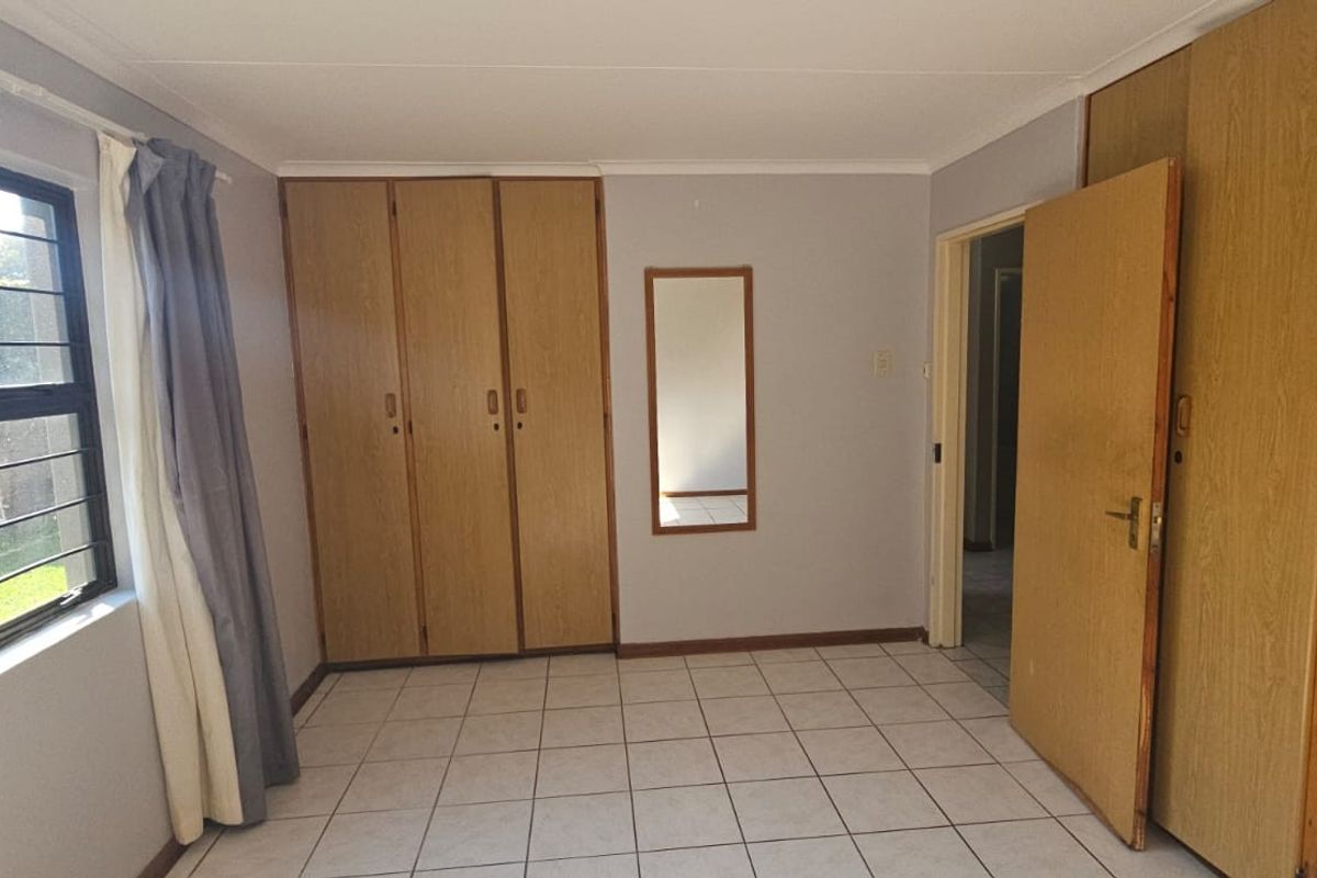 A bedroom featuring tiled flooring, built-in wooden cupboards, and a window with security bars overlooking the garden.