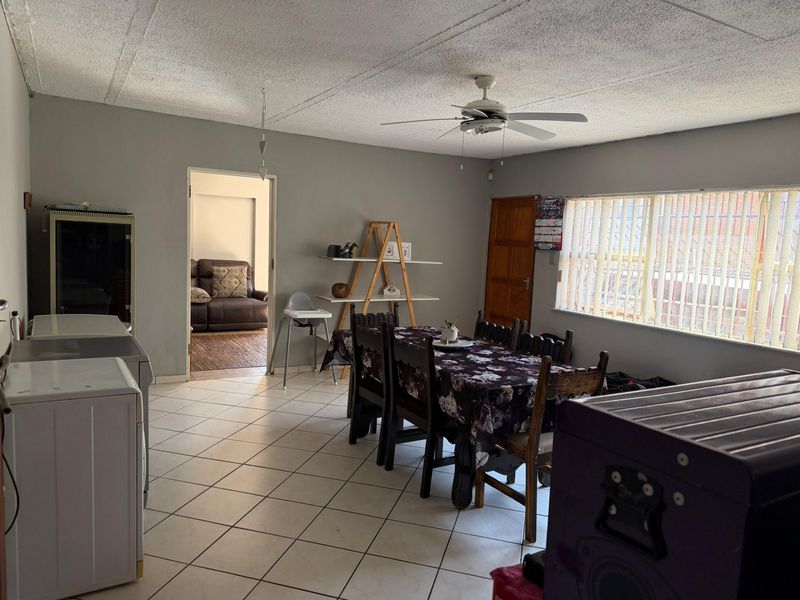 Open plan dining area featuring tiled flooring, a ceiling fan, and large windows with vertical blinds, leading into an adjacent living space.