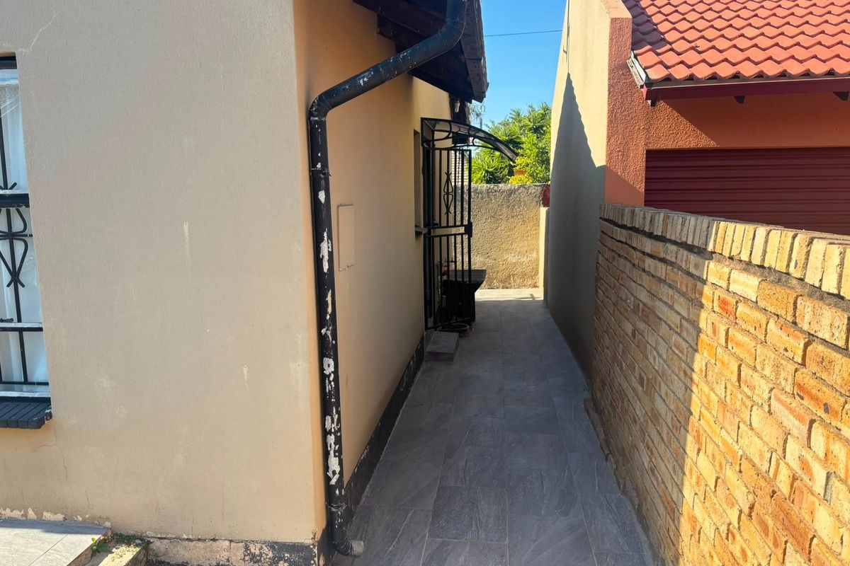 A narrow side passage featuring tiled flooring, a metal security gate, and a downpipe running along the exterior wall of the house.