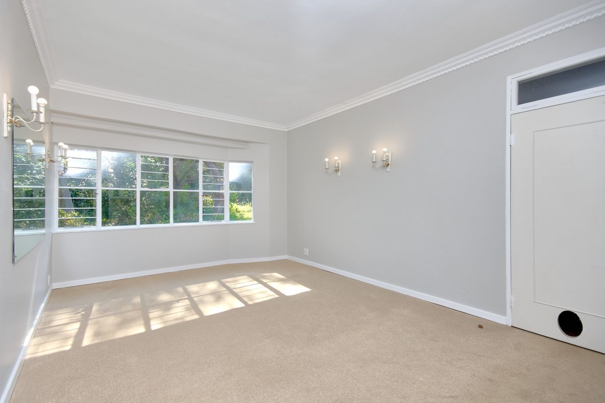 A bright, empty bedroom featuring neutral carpeting, crown molding, wall-mounted light fixtures, and a large window overlooking the garden.