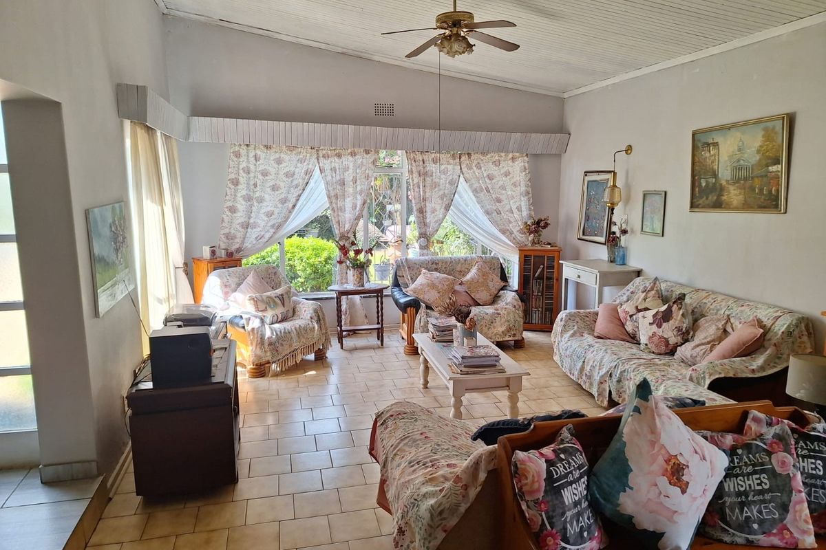 A spacious living room featuring tiled flooring, a vaulted ceiling with a central fan, and large windows providing natural light and garden views.
