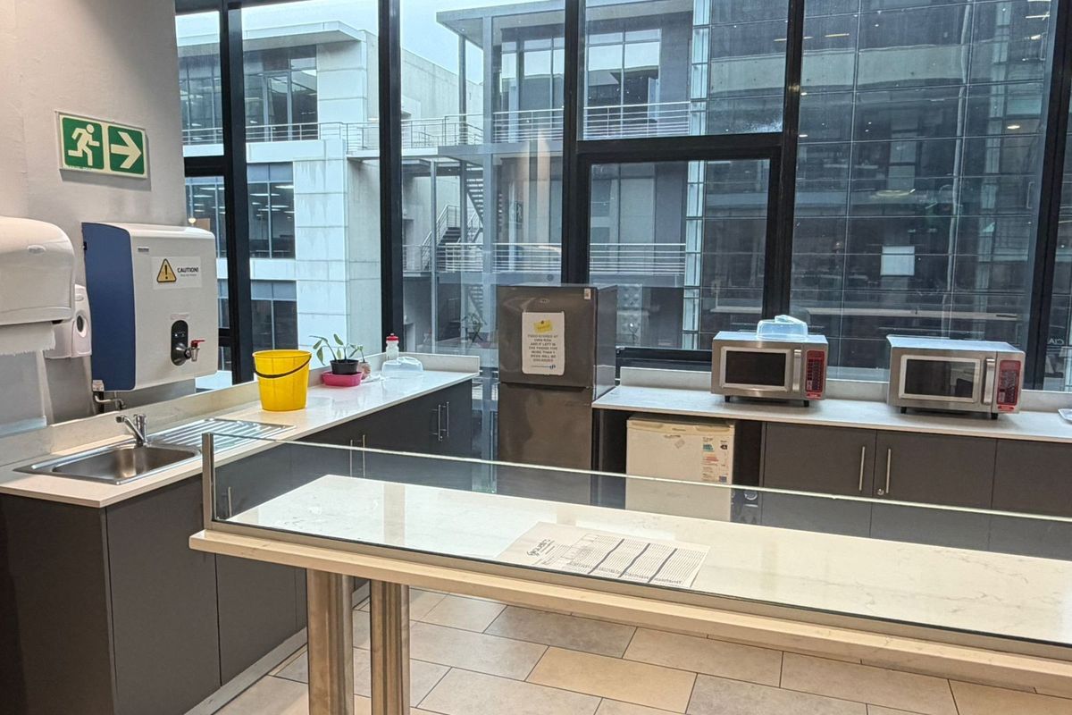 A modern office kitchenette featuring sleek grey cabinetry, stone countertops, a stainless steel sink, and large windows providing ample natural light.