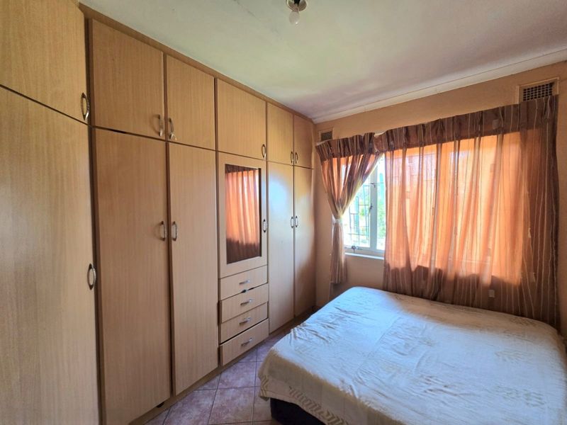 A bedroom featuring extensive built-in wooden cupboards, tiled flooring, and a window with security bars.