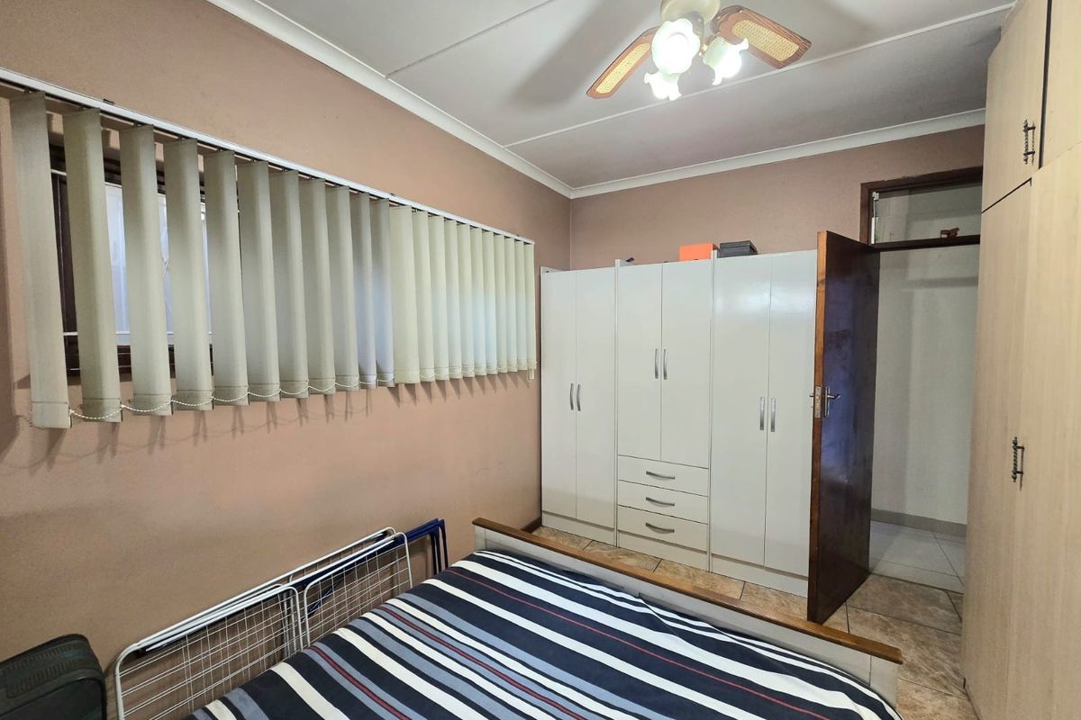 A bedroom featuring built-in cupboards, a ceiling fan, and vertical blinds, with tiled flooring throughout.
