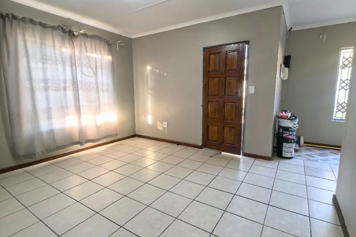 A bright, open-plan living area featuring neutral tiled flooring, a solid timber entrance door, and large windows allowing for ample natural light.
