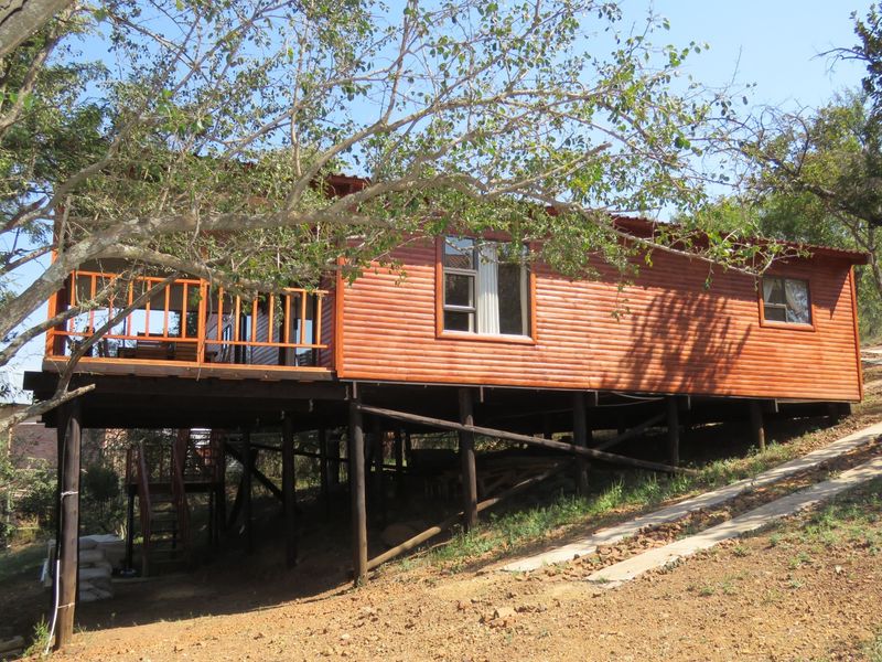 Elevated timber-clad cabin featuring a raised wooden deck and large windows, set on a sloped plot with natural surroundings.