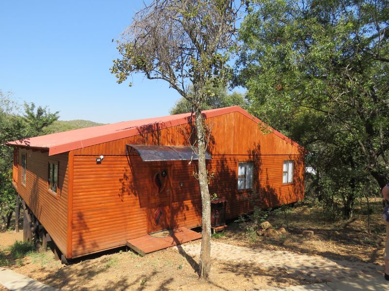 A rustic timber-clad cabin featuring a red metal roof, elevated foundation, and a small covered entrance, set within a natural wooded environment.