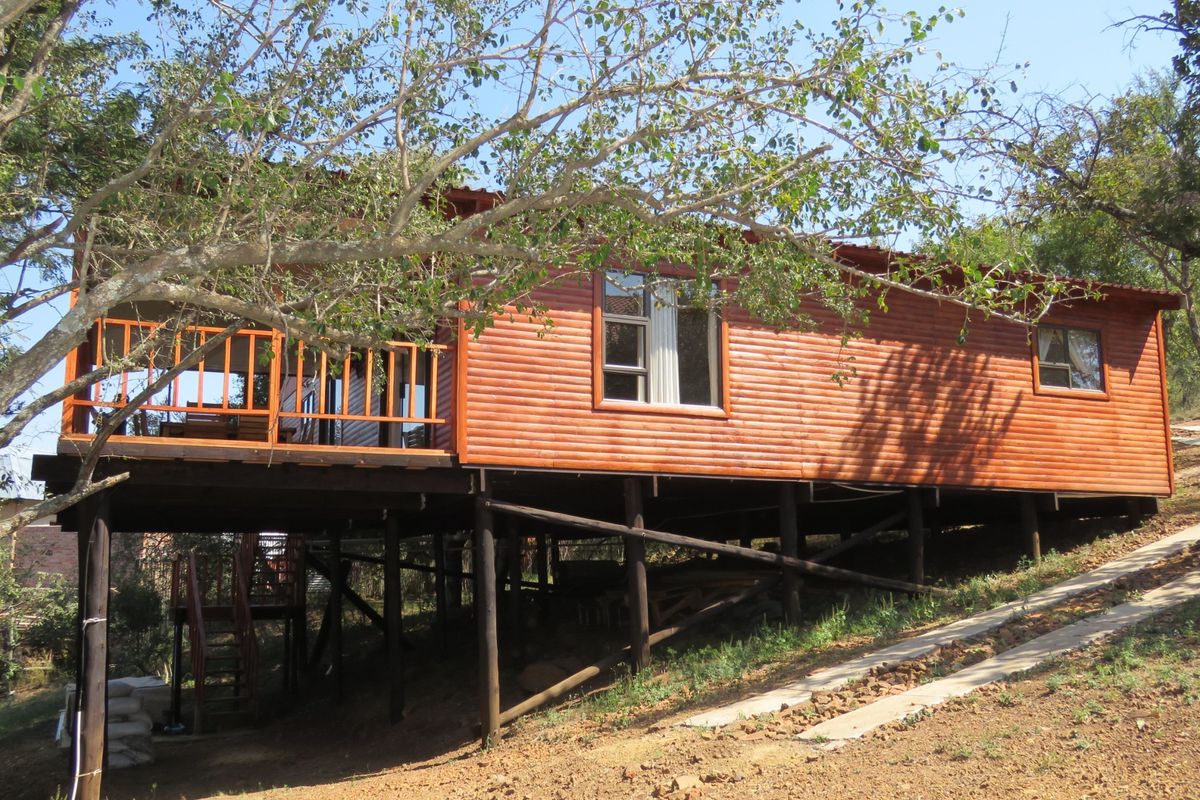Elevated timber-clad cabin featuring a raised wooden deck and large windows, set on a sloped plot with natural surroundings.