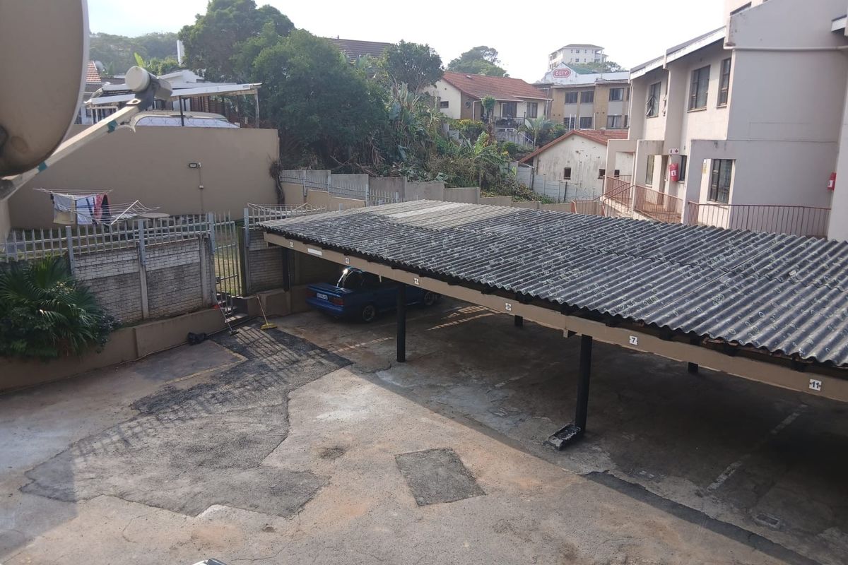 An elevated view of the apartment complex parking area featuring a covered carport structure and paved driveway with perimeter security fencing.