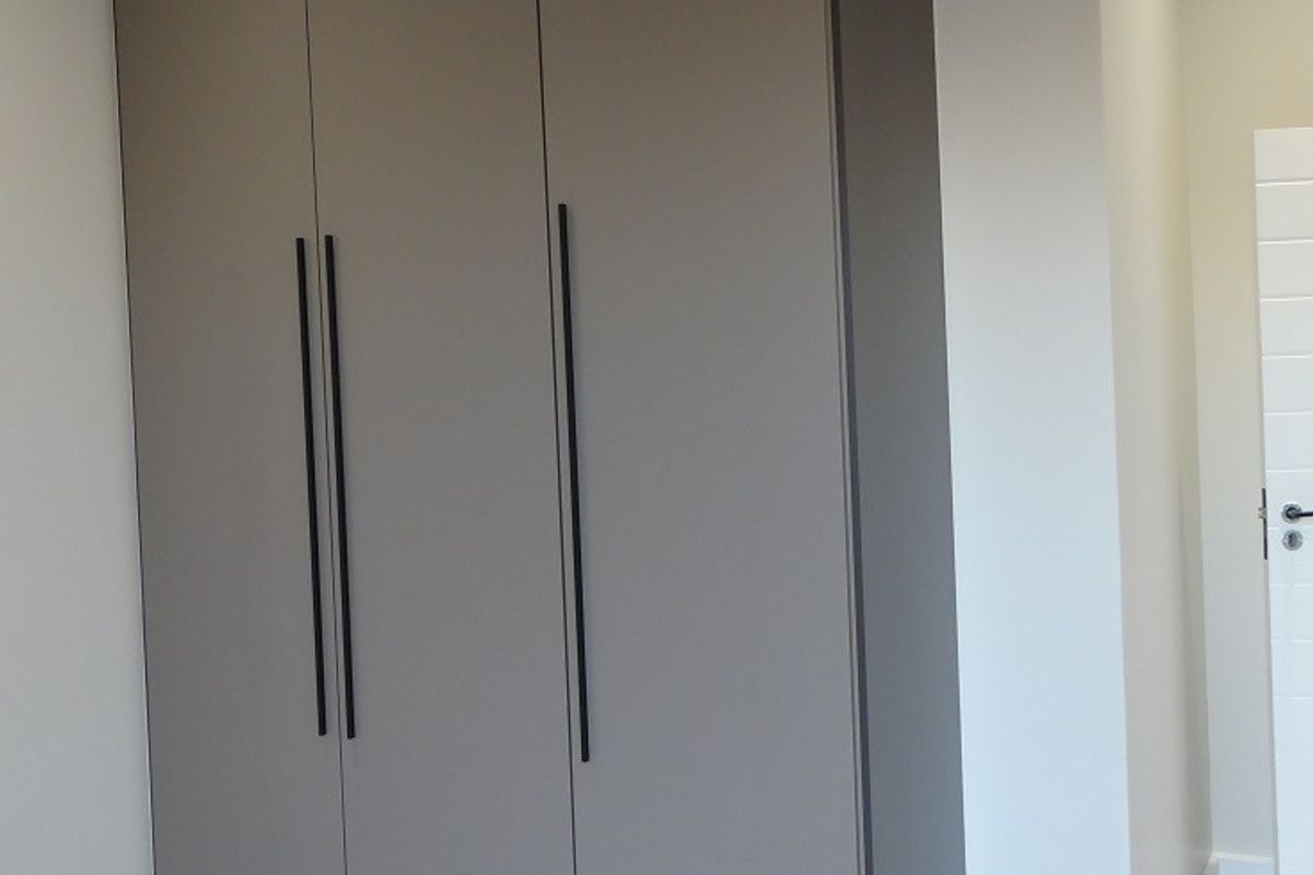 A modern bedroom featuring floor-to-ceiling built-in wardrobes with sleek grey cabinetry, dark trim, and minimalist black handles, set against light wood flooring.