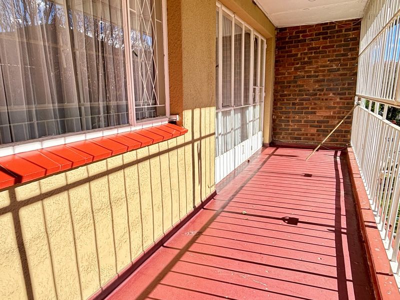 A covered balcony featuring a red floor, exposed brick wall, and security bars, showing signs of wear and requiring maintenance.