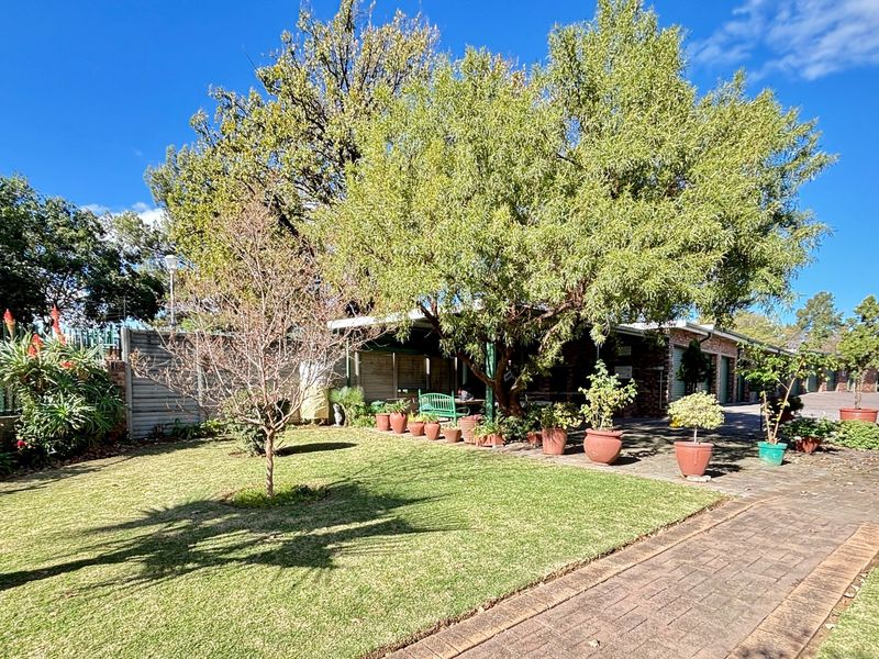 A rear exterior view of the townhouse featuring a well-maintained lawn, paved driveway, and a covered patio area shaded by mature trees.