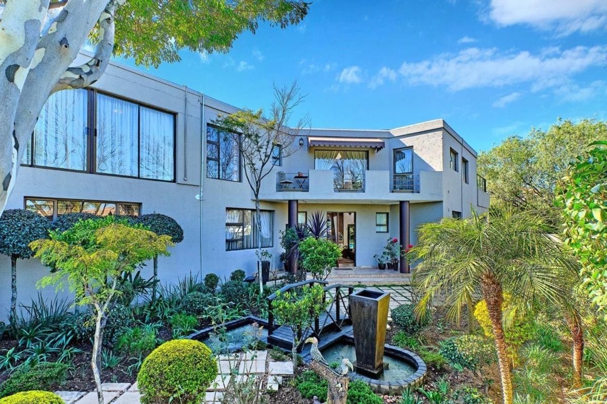 A rear view of a multi-story residence featuring a landscaped garden, a small decorative bridge over a water feature, and an upper-level balcony.