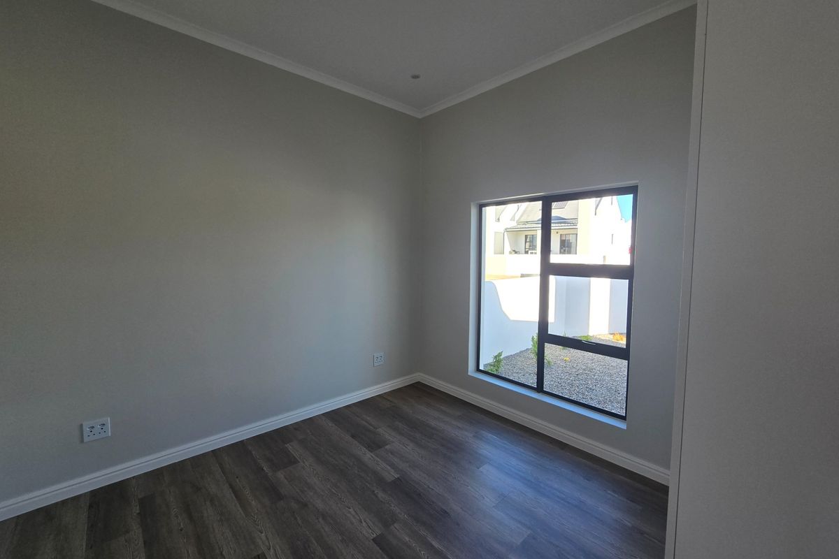 A bright, empty bedroom featuring modern laminate flooring, neutral wall finishes, and a large aluminium-framed window providing natural light.