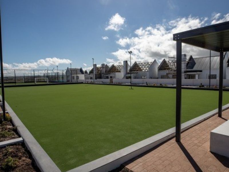 A well-maintained synthetic turf bowling green or sports court featuring concrete edging, a paved walkway, and a modern pergola structure.