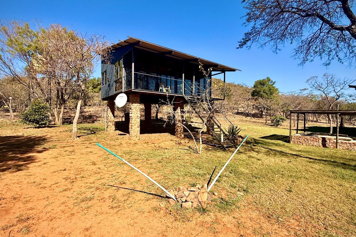 Elevated farm dwelling featuring stone pillar foundations, a wrap-around balcony, and corrugated metal roofing, set within a natural bushveld landscape.