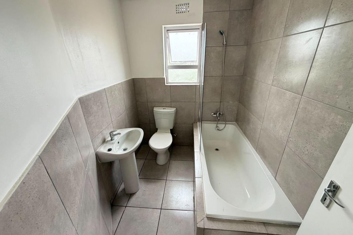 A functional bathroom featuring a pedestal basin, toilet, and a built-in bathtub with a glass shower screen and neutral wall and floor tiling.
