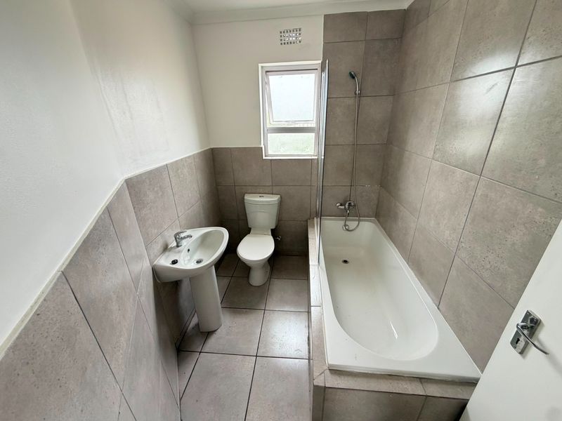 A functional bathroom featuring a pedestal basin, toilet, and a built-in bathtub with a glass shower screen and neutral wall and floor tiling.