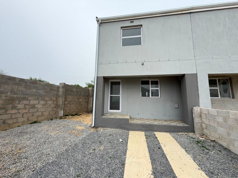 Rear view of a two-story townhouse featuring a paved patio area, gravel yard, and unfinished boundary walls.