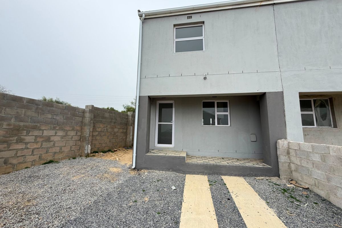 Rear view of a two-story townhouse featuring a paved patio area, gravel yard, and unfinished boundary walls.