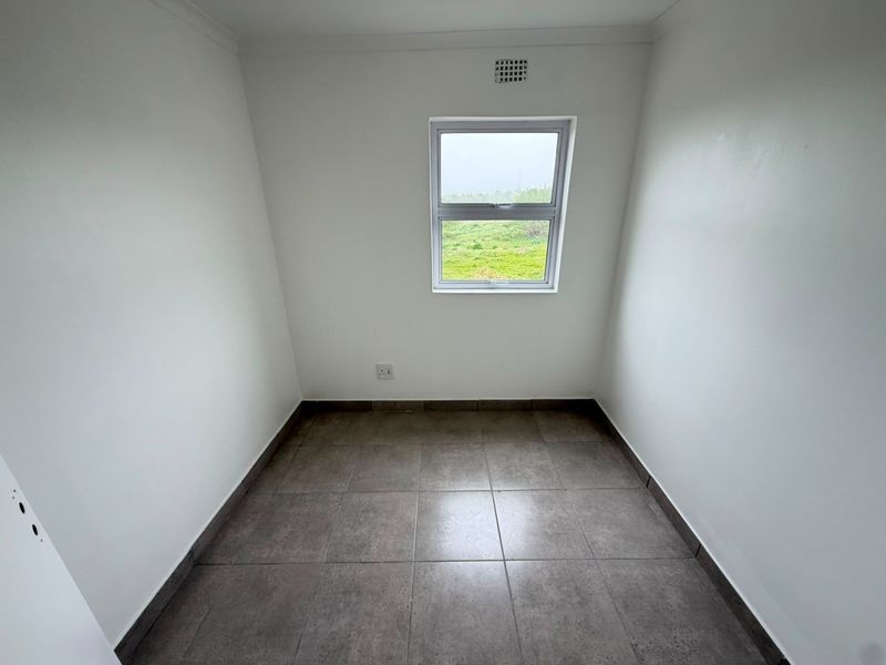 A compact, empty bedroom featuring neutral tiled flooring, white walls, and a single window providing natural light and exterior views.