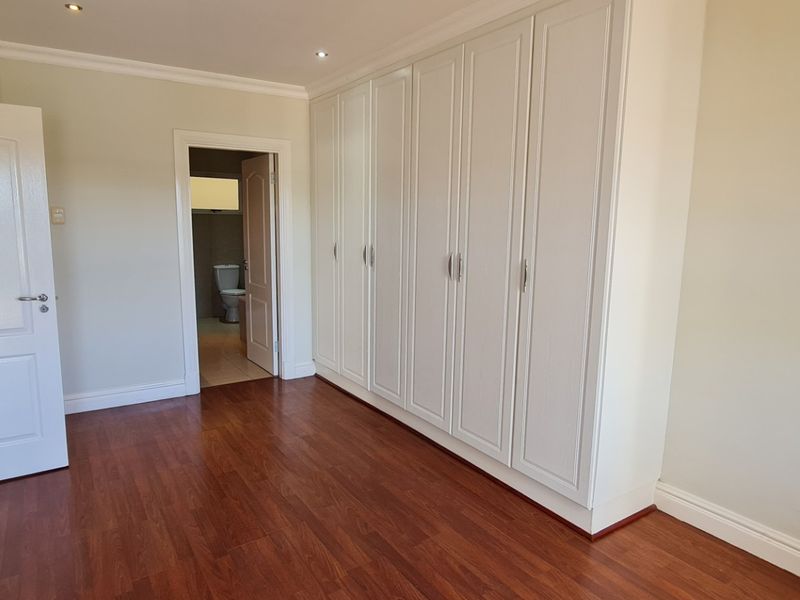 A bright, empty bedroom featuring laminate flooring, extensive built-in cupboards, and recessed downlighting, with an adjoining bathroom visible through the doorway.