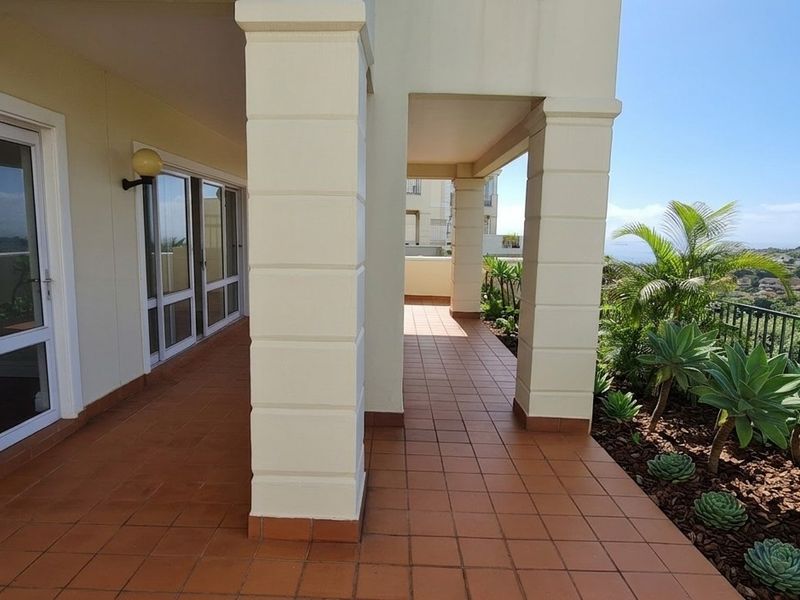 A spacious covered balcony featuring tiled flooring, structural columns, and a metal balustrade, offering scenic views and a landscaped garden border.