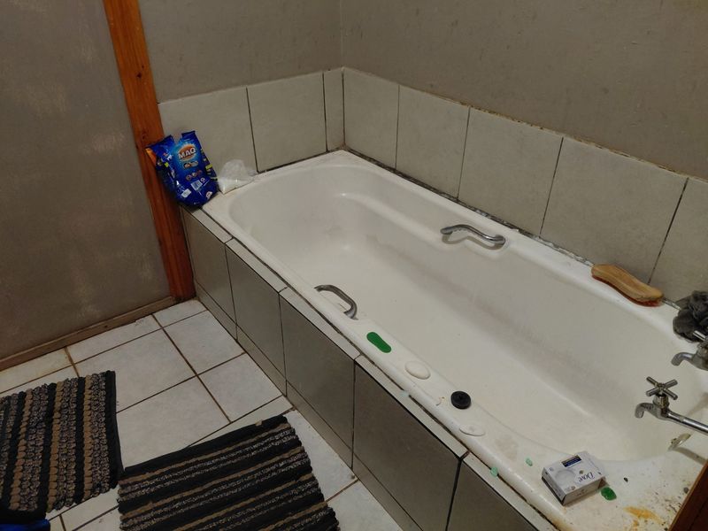 A functional bathroom featuring a built-in bathtub with tiled surround and chrome tapware, requiring cosmetic updates to finishes and grout.
