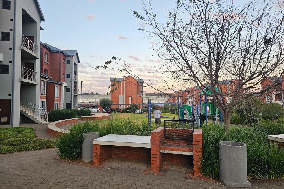 Communal outdoor area featuring a brick-built barbecue station, paved walkways, landscaped garden beds, and a children's playground set within an apartment complex.