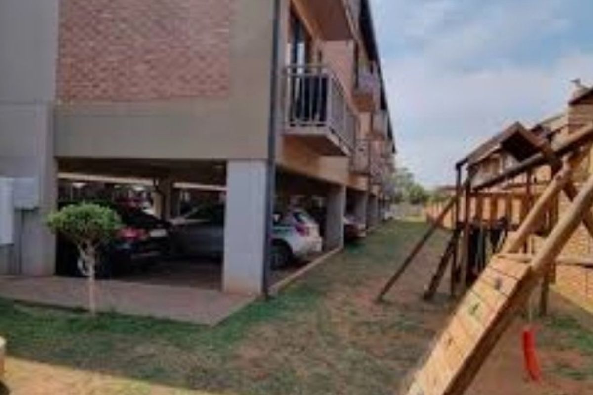 Exterior view of the apartment complex featuring covered ground-level parking bays and a communal outdoor play area with a wooden climbing frame.