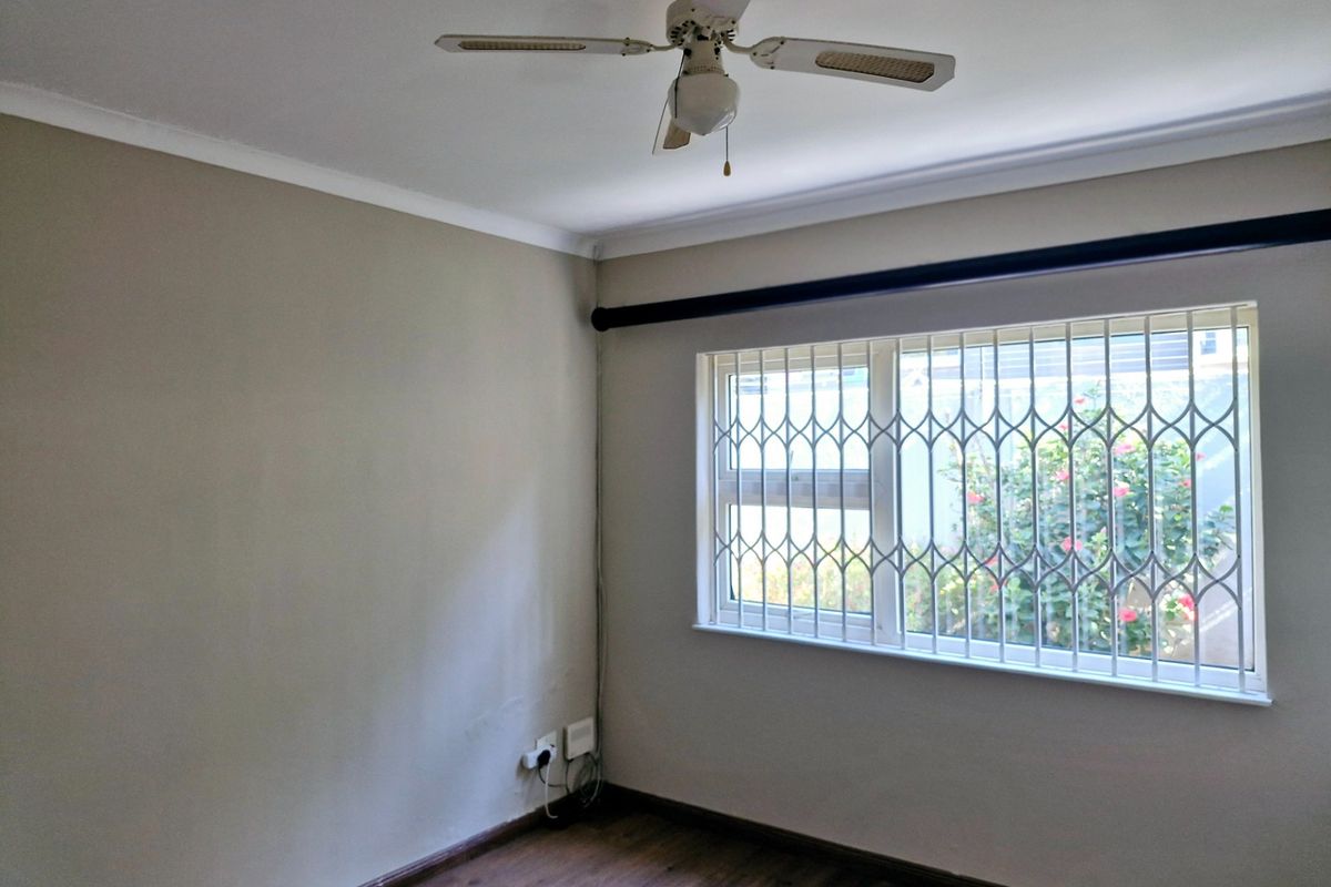 A bright, empty bedroom featuring wood-look flooring, a ceiling fan, and a window fitted with security bars.