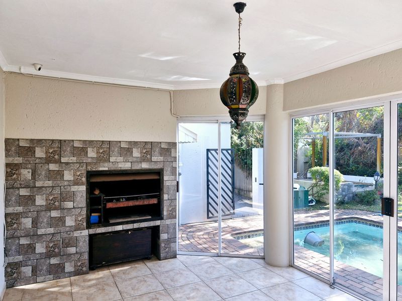 Covered patio area featuring a built-in stone-clad braai, tiled flooring, and sliding glass doors providing direct access to the swimming pool and garden.