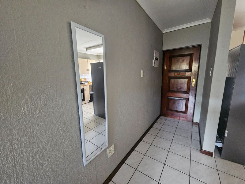 A functional apartment hallway featuring tiled flooring, a solid wood entrance door, and a wall-mounted mirror, providing access to the adjacent kitchen area.