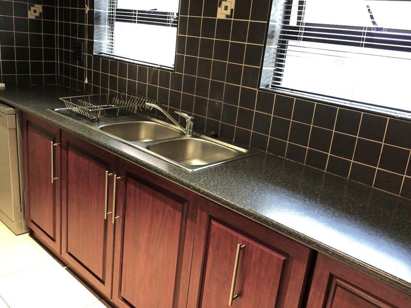 A functional kitchen area featuring dark tiled splashbacks, laminate countertops, and built-in wooden cabinetry with a double bowl stainless steel sink.