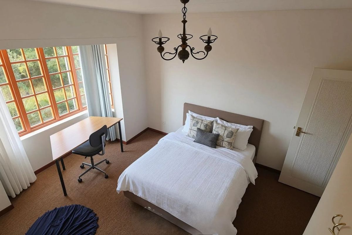 A well-lit bedroom featuring carpeted flooring, a classic wrought iron chandelier, and a large multi-pane window providing natural light.